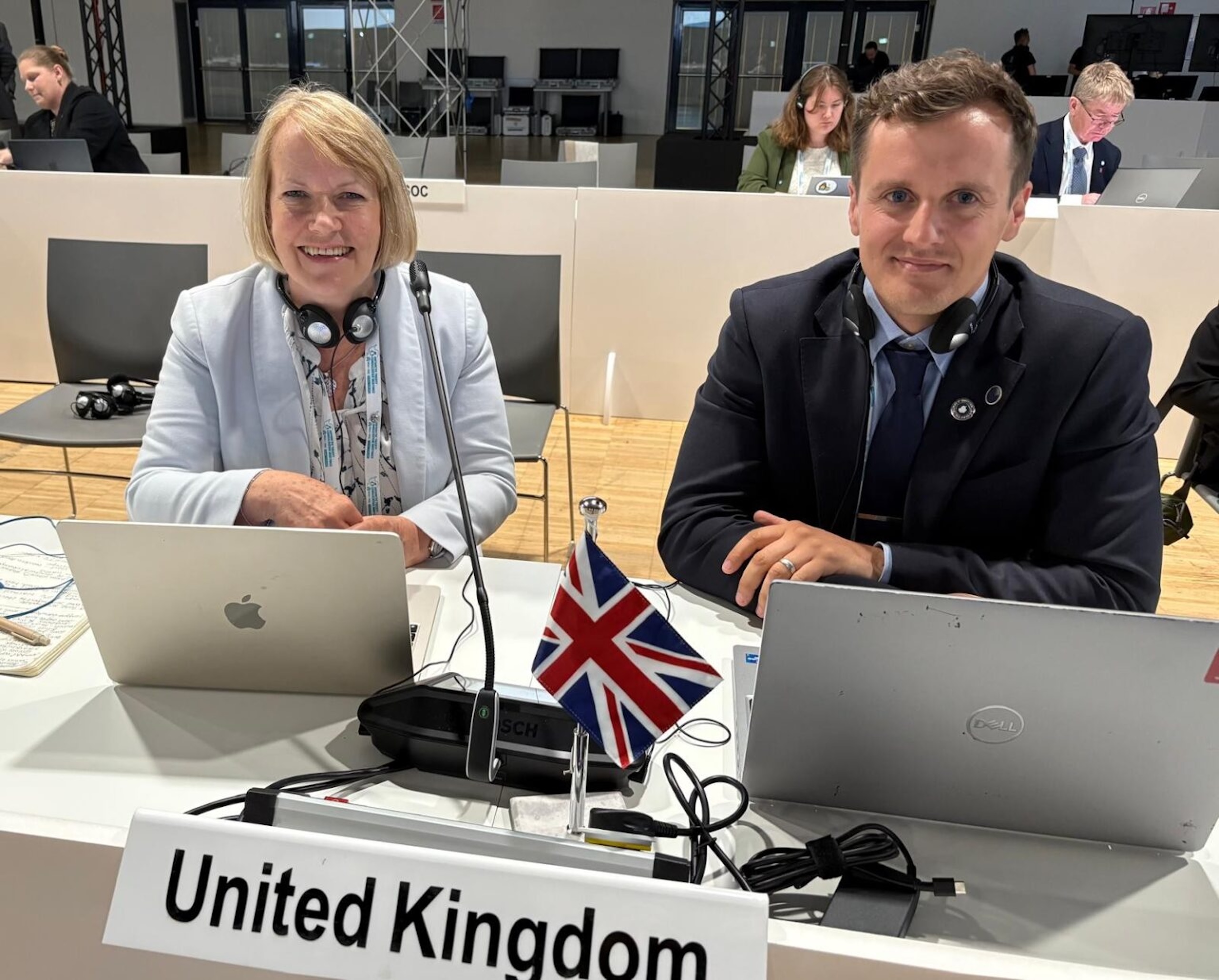 Jane and Ollie sit in the ATCM conference hall side by side at a desk. They both have laptops in front of them, plus a little UK flag and a sign saying 'United Kingdom'.
