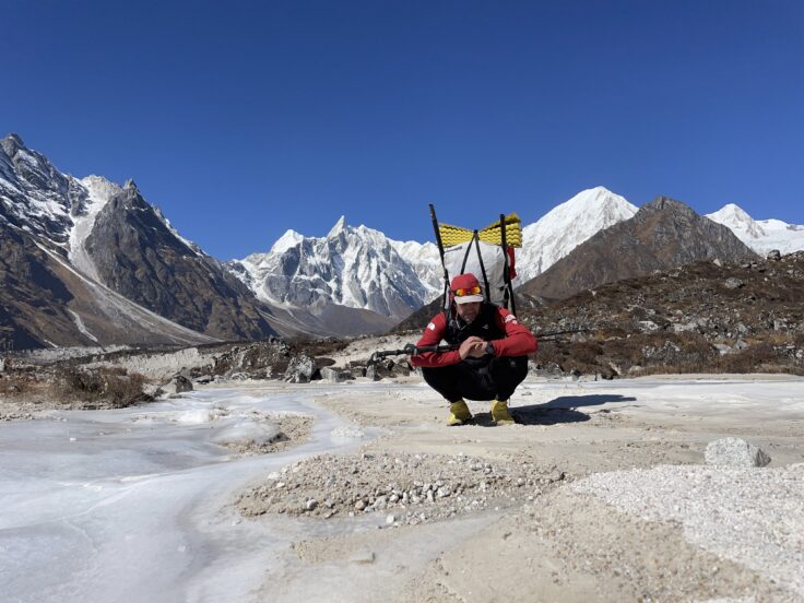 A person crouches to rest in the final moments of a hike to Rara Lake in the Himalaya. They have large a backpack on, in the distance snowy mountains rise above the hilly landscape.