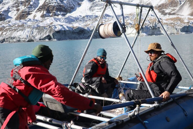A group are sitting on a custom boat rig on a mountainous Himalayan lake. They are wearing life jackets, cold weather gear and sunglasses. The rig above them has a real of rope designed to pull back up sampling instruments from the lake bed.