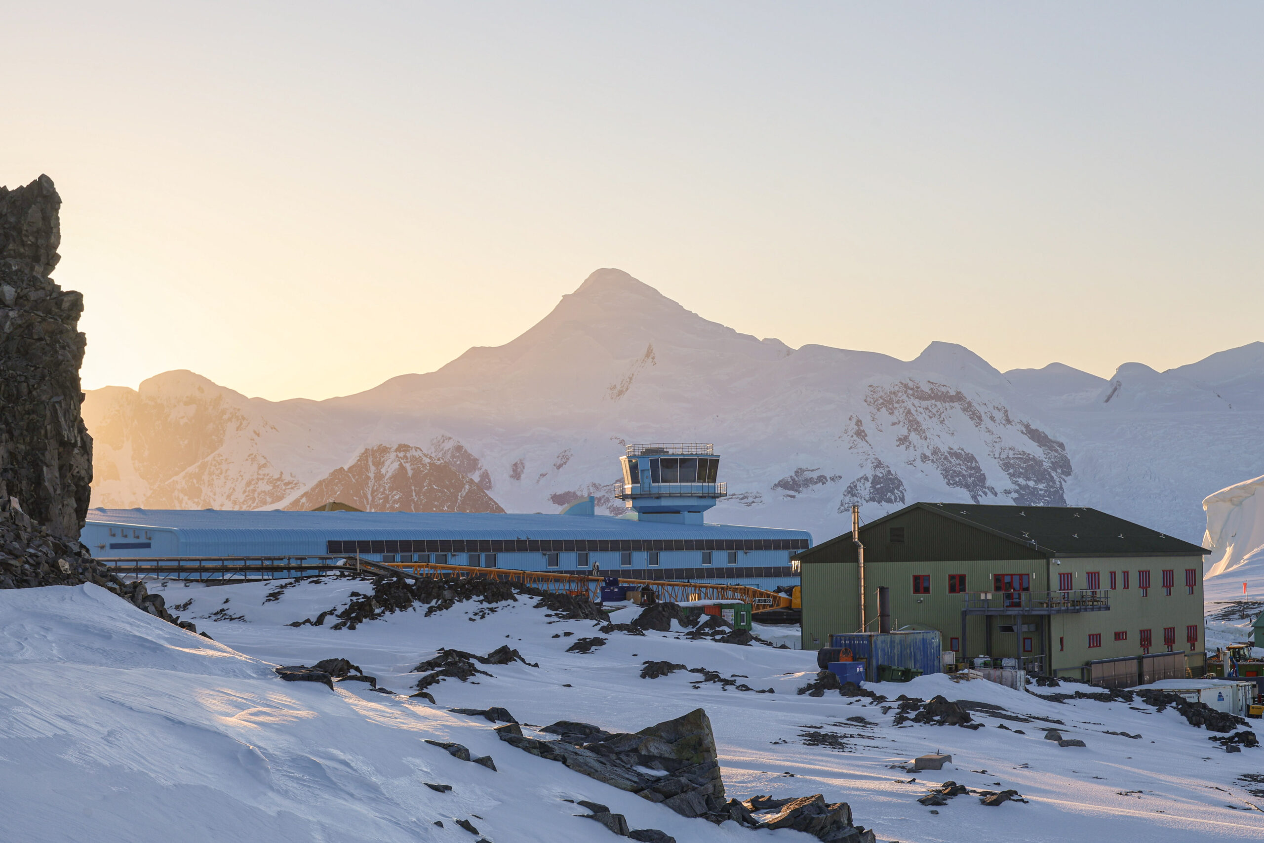  A view over Rothera Research Station with the sun low on the horizon. There is a large blue building in the background and a smaller green building in front of it. 