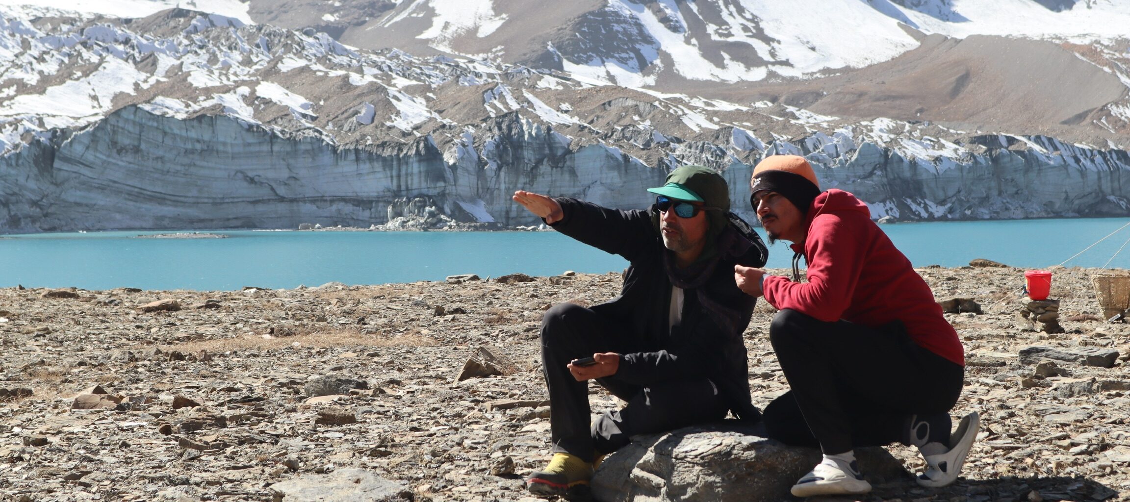 Two people kneel on the rocky outcrop next to a bright blue lake, behind which huge glaciers and mountains rise out of frame. They are looking and pointing into the distance.