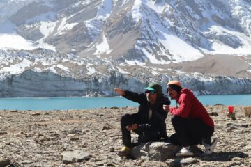 Two people kneel on the rocky outcrop next to a bright blue lake, behind which huge glaciers and mountains rise out of frame. They are looking and pointing into the distance.