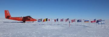 A propellor areoplane next to the flags of the Antarctic Treaty nations, behind the south pole marker