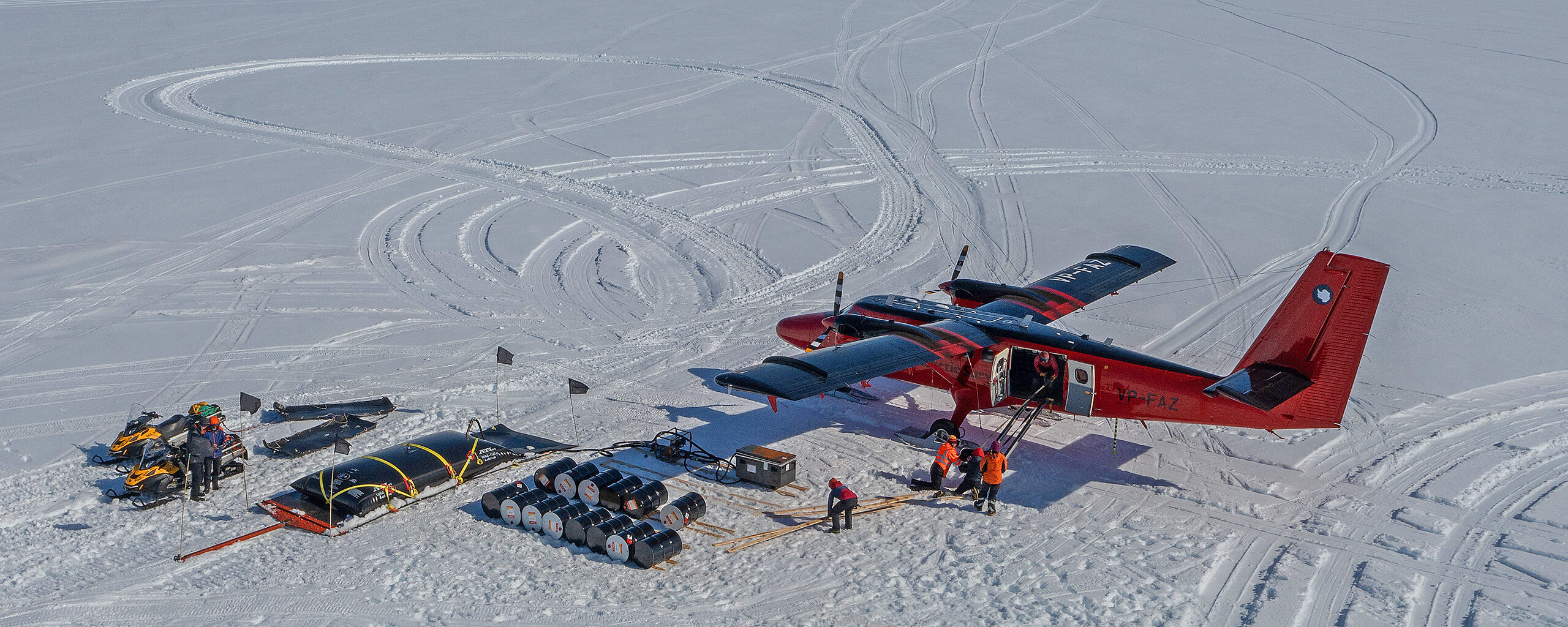 A red Twin Otter aircraft loading cargo including fuel drums at Gromits Creek, Antarctica.